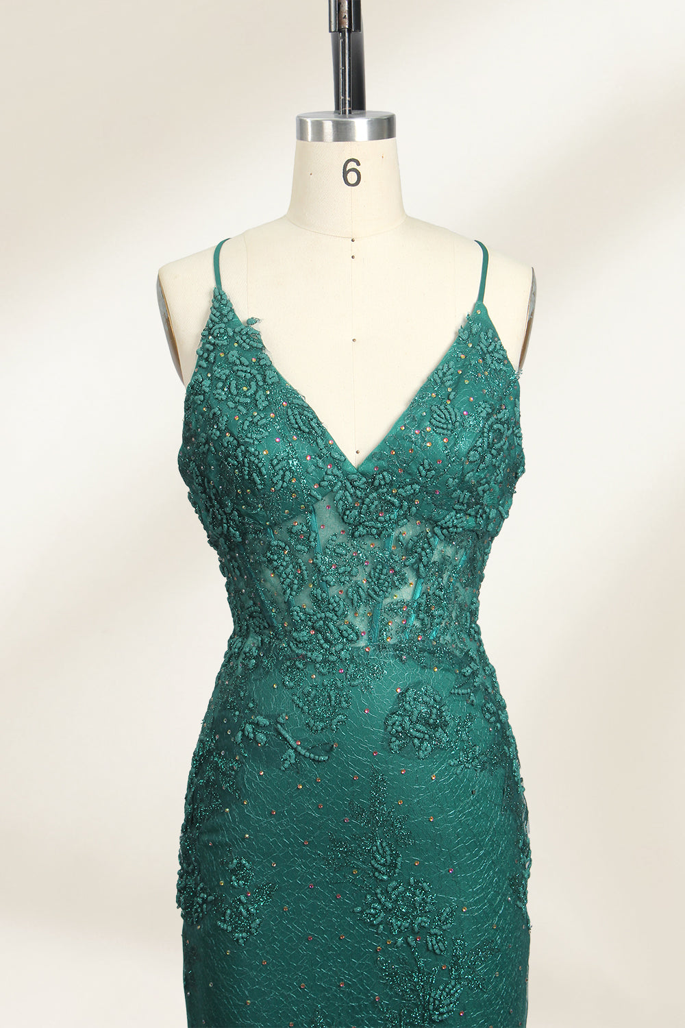 Photograph of a dark green, floor-length evening gown displayed on a white dress form. The gown features a deep V-neckline with thin spaghetti straps, a fitted bodice adorned with intricate lace detailing, and a flowing, slightly flared skirt. The dress showcases a rich, textured fabric with a shimmering effect, accentuating the luxurious and elegant style. The background is a plain, off-white color, ensuring the dress remains the focal point.02
