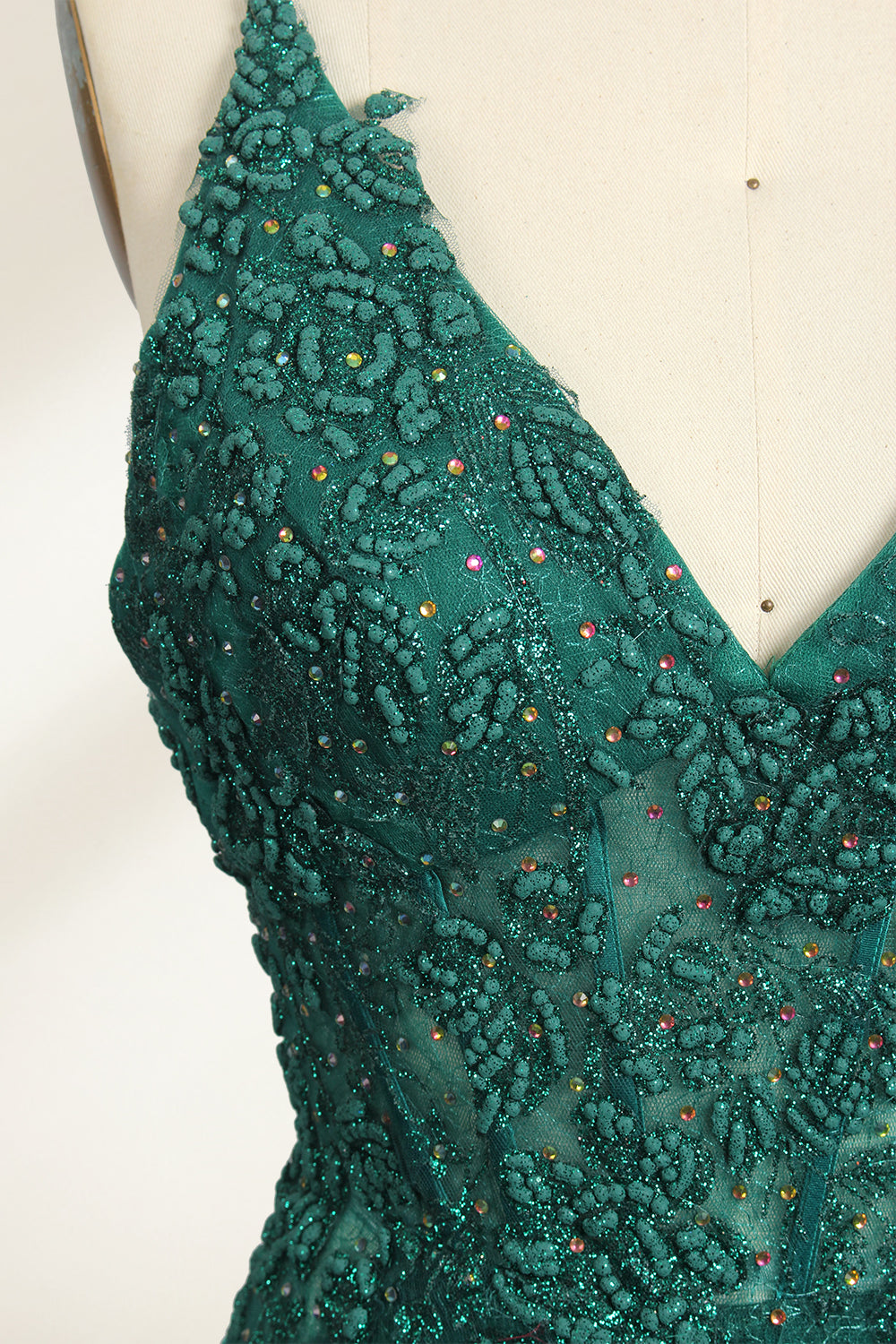 Photograph of a dark green, floor-length evening gown displayed on a white dress form. The gown features a deep V-neckline with thin spaghetti straps, a fitted bodice adorned with intricate lace detailing, and a flowing, slightly flared skirt. The dress showcases a rich, textured fabric with a shimmering effect, accentuating the luxurious and elegant style. The background is a plain, off-white color, ensuring the dress remains the focal point.06