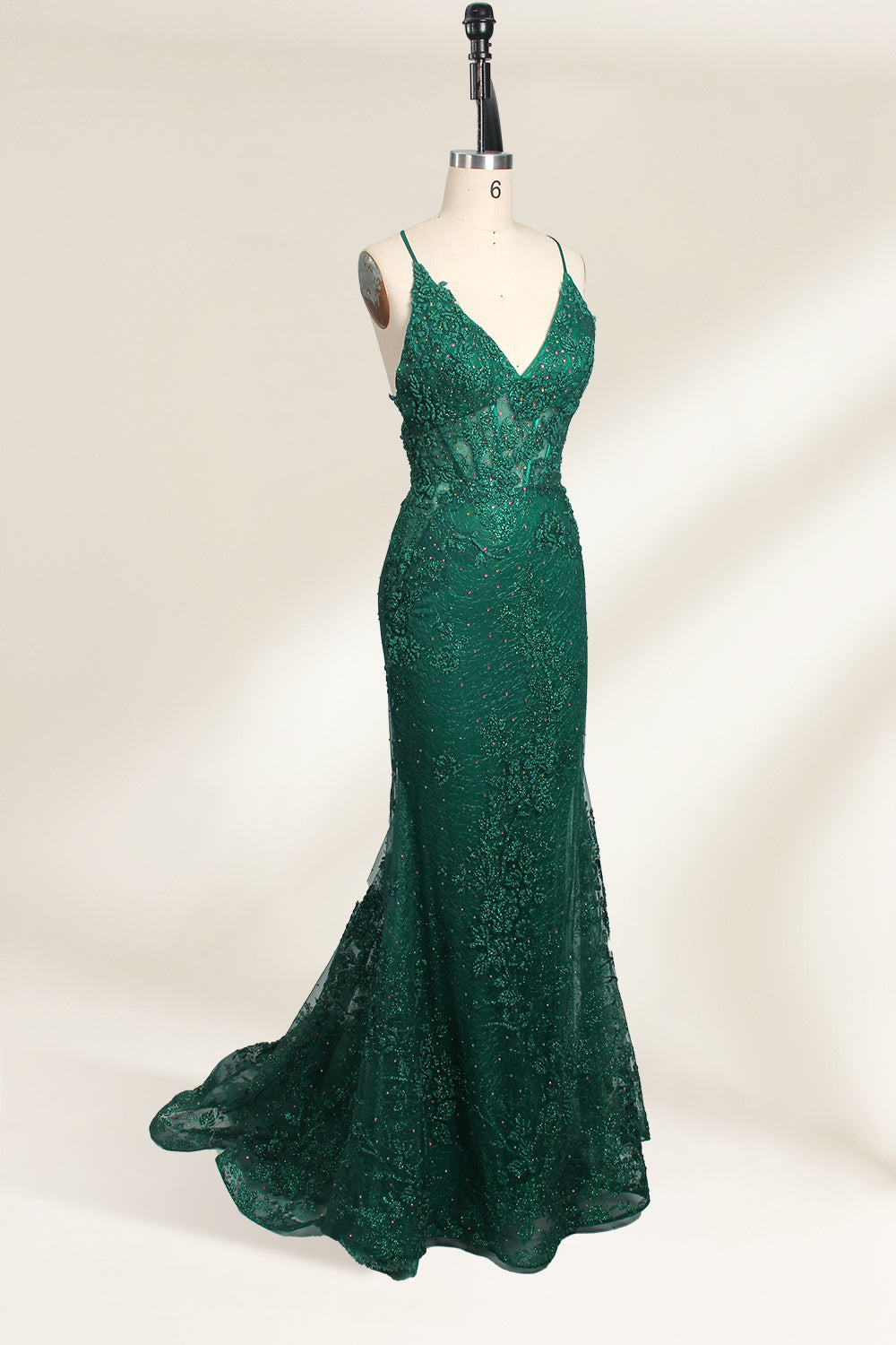 Photograph of a dark green, floor-length evening gown displayed on a white dress form. The gown features a deep V-neckline with thin spaghetti straps, a fitted bodice adorned with intricate lace detailing, and a flowing, slightly flared skirt. The dress showcases a rich, textured fabric with a shimmering effect, accentuating the luxurious and elegant style. The background is a plain, off-white color, ensuring the dress remains the focal point.04