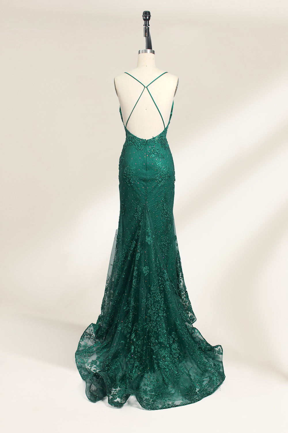 Photograph of a dark green, floor-length evening gown displayed on a white dress form. The gown features a deep V-neckline with thin spaghetti straps, a fitted bodice adorned with intricate lace detailing, and a flowing, slightly flared skirt. The dress showcases a rich, textured fabric with a shimmering effect, accentuating the luxurious and elegant style. The background is a plain, off-white color, ensuring the dress remains the focal point.03
