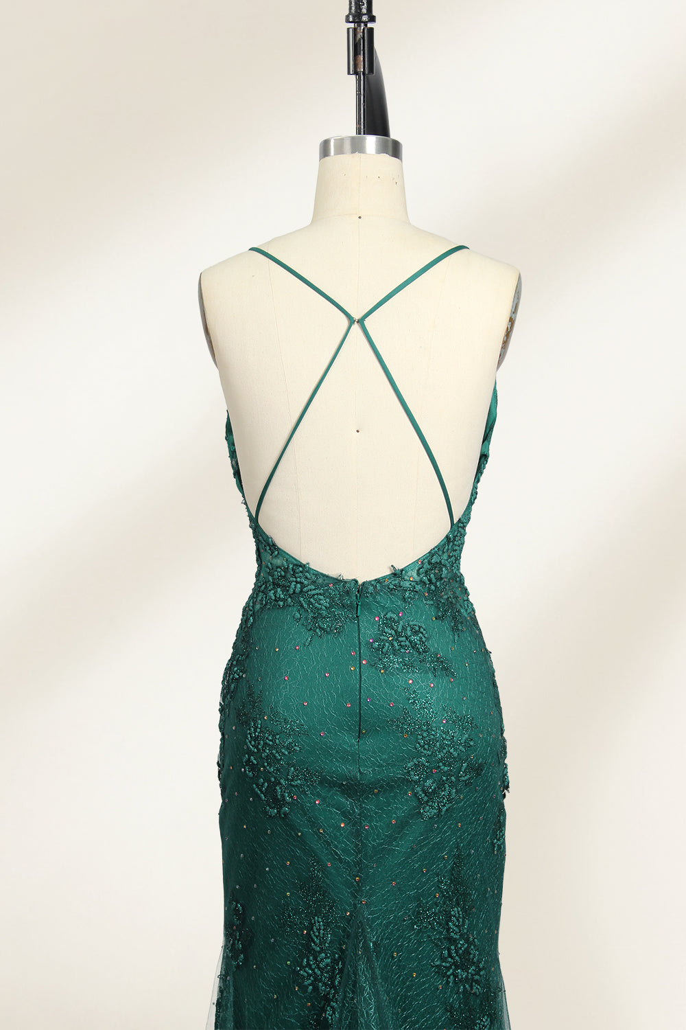 Photograph of a dark green, floor-length evening gown displayed on a white dress form. The gown features a deep V-neckline with thin spaghetti straps, a fitted bodice adorned with intricate lace detailing, and a flowing, slightly flared skirt. The dress showcases a rich, textured fabric with a shimmering effect, accentuating the luxurious and elegant style. The background is a plain, off-white color, ensuring the dress remains the focal point.05