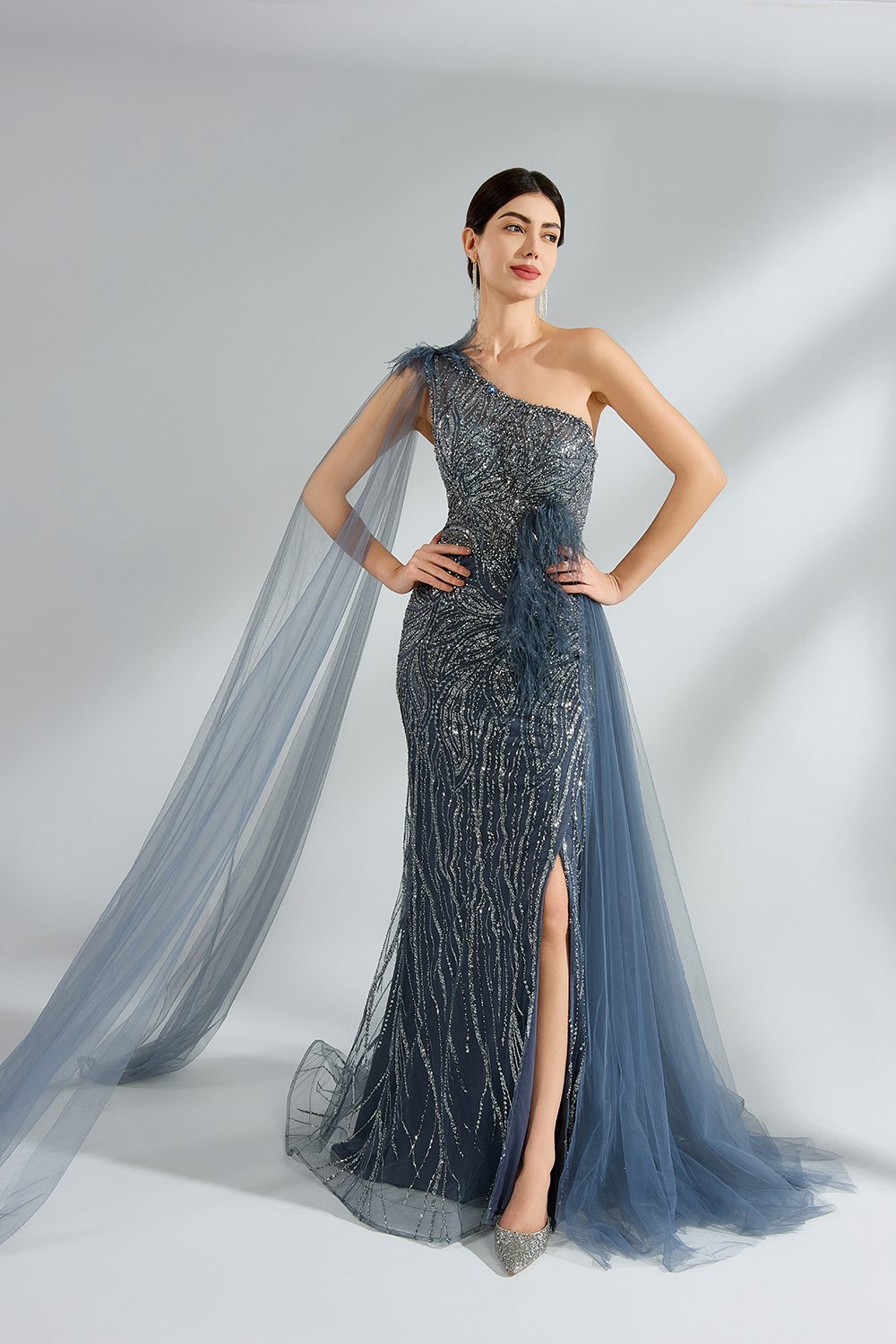 This image features a woman in a glamorous, one-shoulder gown with intricate beadwork and a flowing tulle train. The composition highlights elegance and sophistication, with a minimalist background emphasizing the dress's luxurious texture and shimmering details.