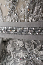 Displayed on a mannequin is a silver - gray evening gown. It features a sheer neckline with fringe details, intricate lace embroidery on the bodice, and a flowing tulle overlay. The elegant design creates a sophisticated and glamorous look, perfect for formal events.6