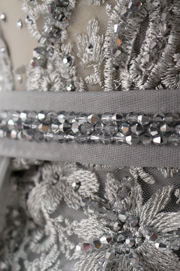 Displayed on a mannequin is a silver - gray evening gown. It features a sheer neckline with fringe details, intricate lace embroidery on the bodice, and a flowing tulle overlay. The elegant design creates a sophisticated and glamorous look, perfect for formal events.6