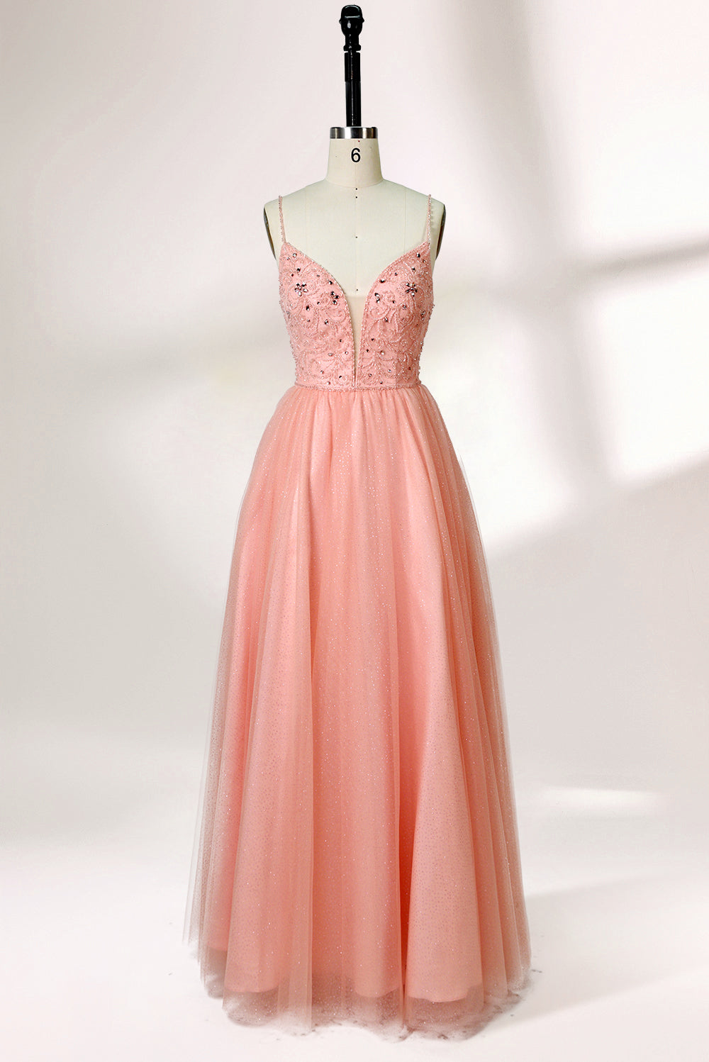 Photograph of a mannequin wearing an elegant, floor-length, blush pink tulle gown with a fitted bodice adorned with sequin embellishments and thin spaghetti straps. The gown has a sweetheart neckline and a full skirt with a subtle sparkle, creating a soft, ethereal effect. The background is a plain white studio setting with soft lighting casting gentle shadows.01