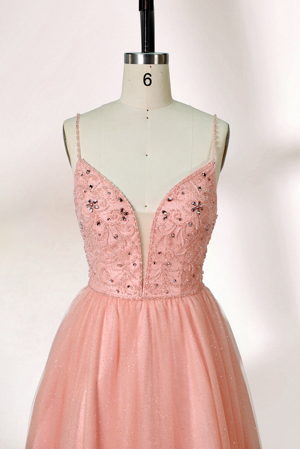Photograph of a mannequin wearing an elegant, floor-length, blush pink tulle gown with a fitted bodice adorned with sequin embellishments and thin spaghetti straps. The gown has a sweetheart neckline and a full skirt with a subtle sparkle, creating a soft, ethereal effect. The background is a plain white studio setting with soft lighting casting gentle shadows.03