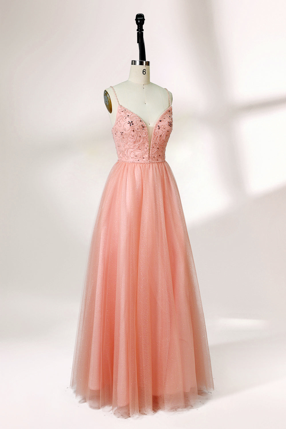 Photograph of a mannequin wearing an elegant, floor-length, blush pink tulle gown with a fitted bodice adorned with sequin embellishments and thin spaghetti straps. The gown has a sweetheart neckline and a full skirt with a subtle sparkle, creating a soft, ethereal effect. The background is a plain white studio setting with soft lighting casting gentle shadows.04