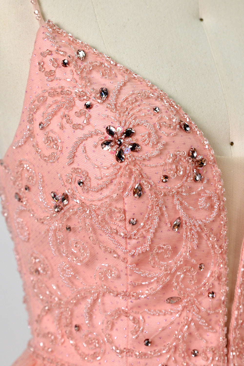 Photograph of a mannequin wearing an elegant, floor-length, blush pink tulle gown with a fitted bodice adorned with sequin embellishments and thin spaghetti straps. The gown has a sweetheart neckline and a full skirt with a subtle sparkle, creating a soft, ethereal effect. The background is a plain white studio setting with soft lighting casting gentle shadows.06