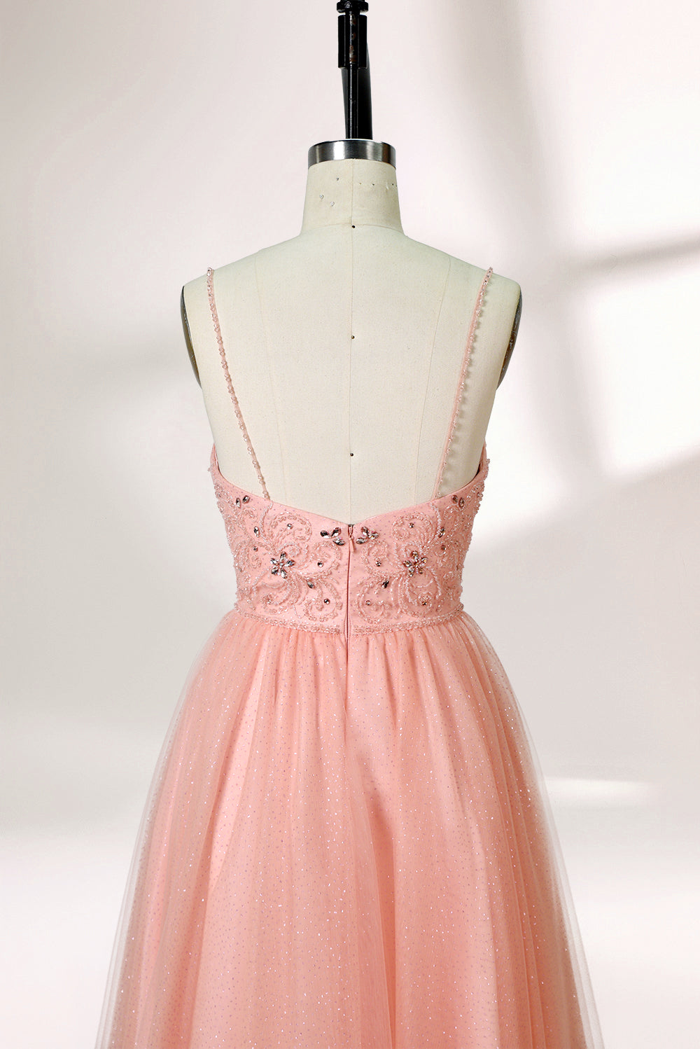 Photograph of a mannequin wearing an elegant, floor-length, blush pink tulle gown with a fitted bodice adorned with sequin embellishments and thin spaghetti straps. The gown has a sweetheart neckline and a full skirt with a subtle sparkle, creating a soft, ethereal effect. The background is a plain white studio setting with soft lighting casting gentle shadows.05