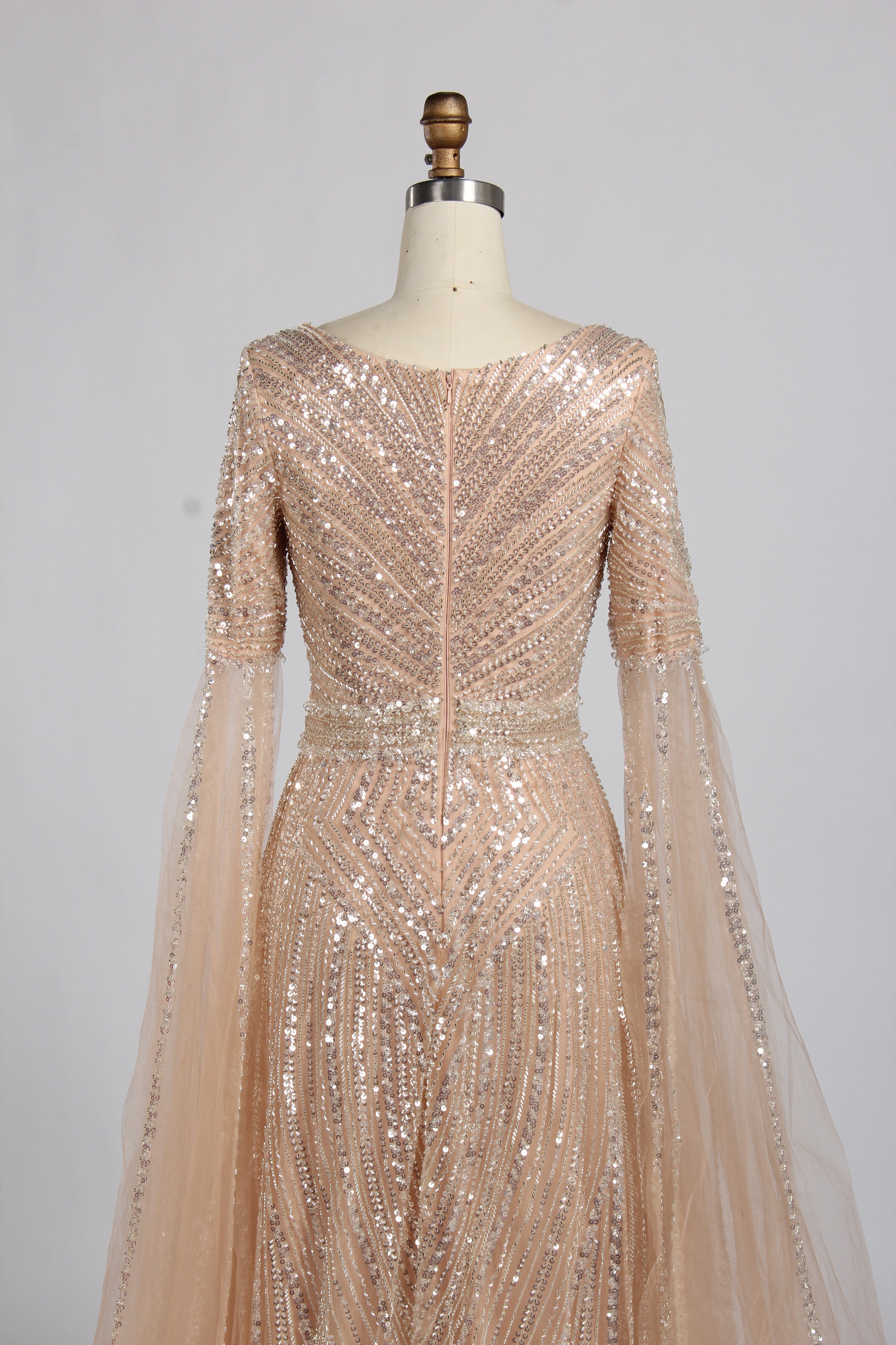 Luxury champagne-colored A-line evening gown on a mannequin, featuring dramatic floor-length cape sleeves, a V-neckline, and shimmering hand-beaded embellishments throughout the tulle fabric.