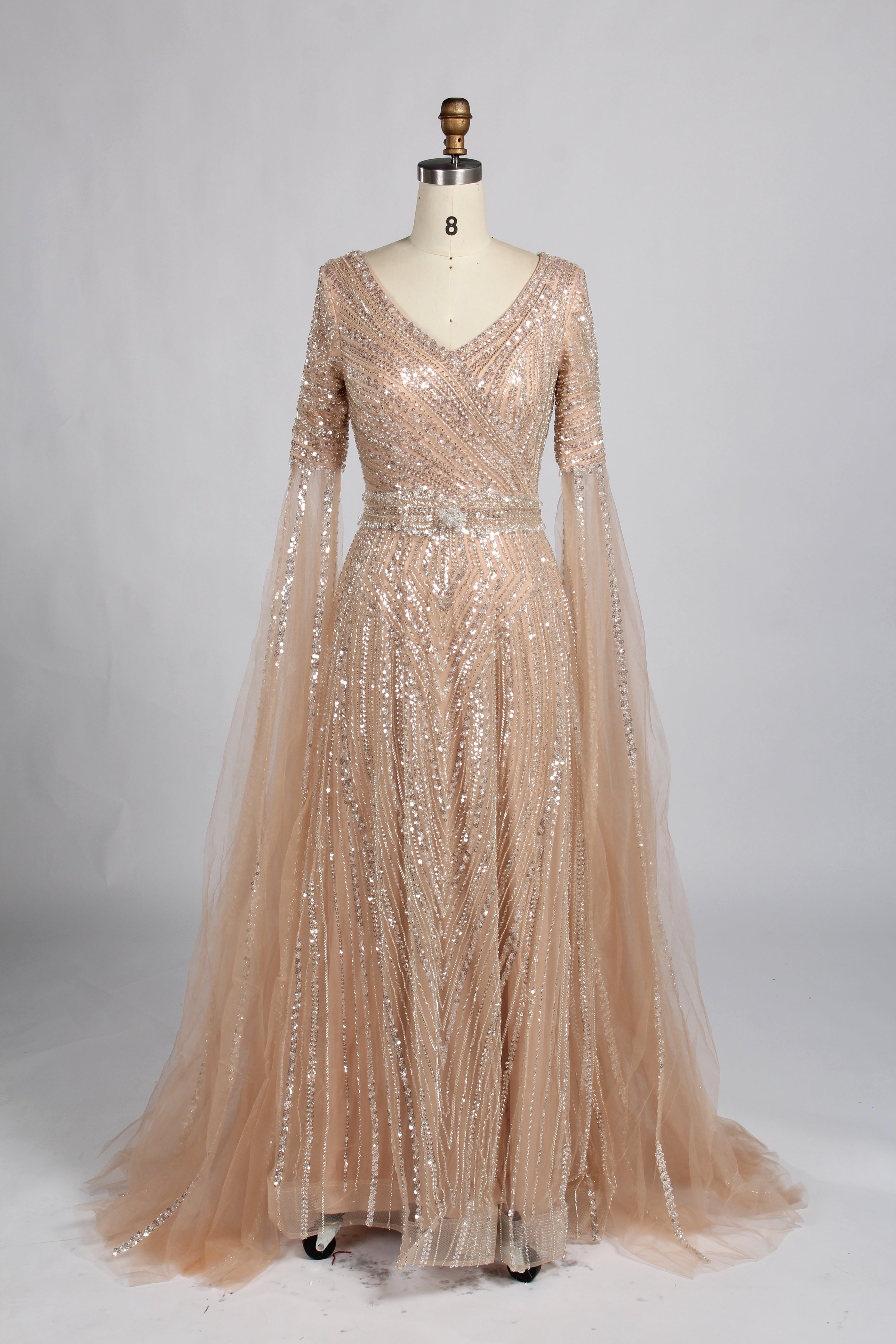 Luxury champagne-colored A-line evening gown on a mannequin, featuring dramatic floor-length cape sleeves, a V-neckline, and shimmering hand-beaded embellishments throughout the tulle fabric.